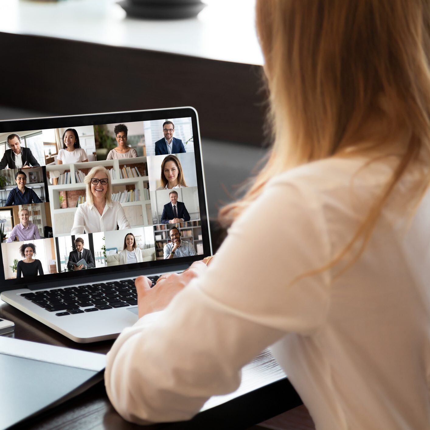 View over businesswoman shoulder sit at workplace desk looks at pc screen during group videocall with different age and ethnicity colleagues using application and webcam, distant communication concept